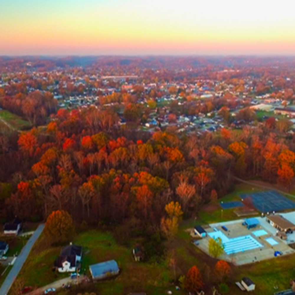 A vibrant aerial view of Flatwoods, Kentucky, during autumn, showcasing a dense canopy of colorful fall foliage, residential neighborhoods, and a public pool area under a clear sky at sunset.