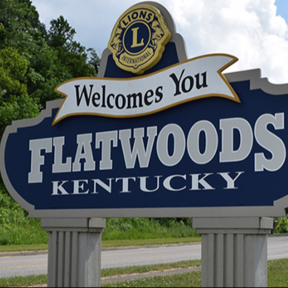 Roadside welcome sign reading “Flatwoods, Kentucky Welcomes You” sponsored by the Lions Club, with a blue and white color scheme surrounded by greenery under a sunny sky.