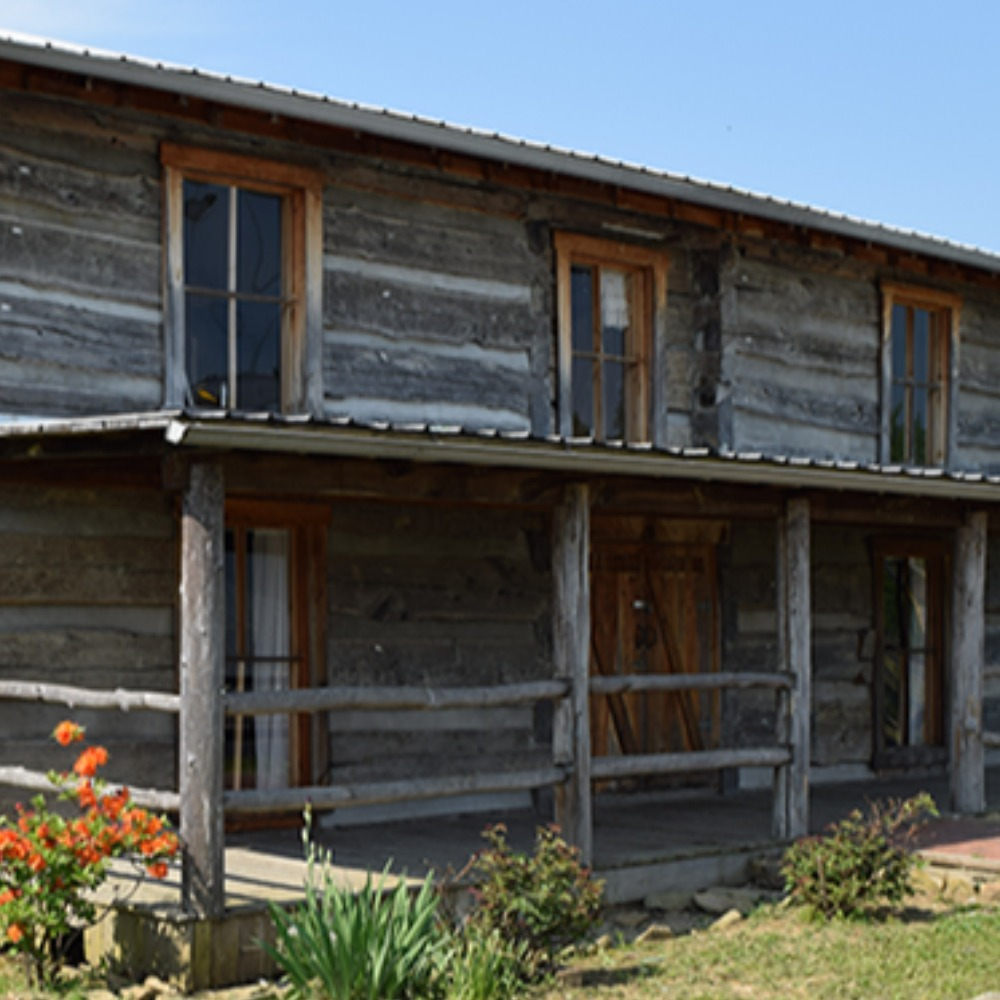 Two-story log cabin known as the Gilley Cabin in Flatwoods, Kentucky, featuring rustic wooden siding, a covered front porch, and blooming flowers in a small garden in front.