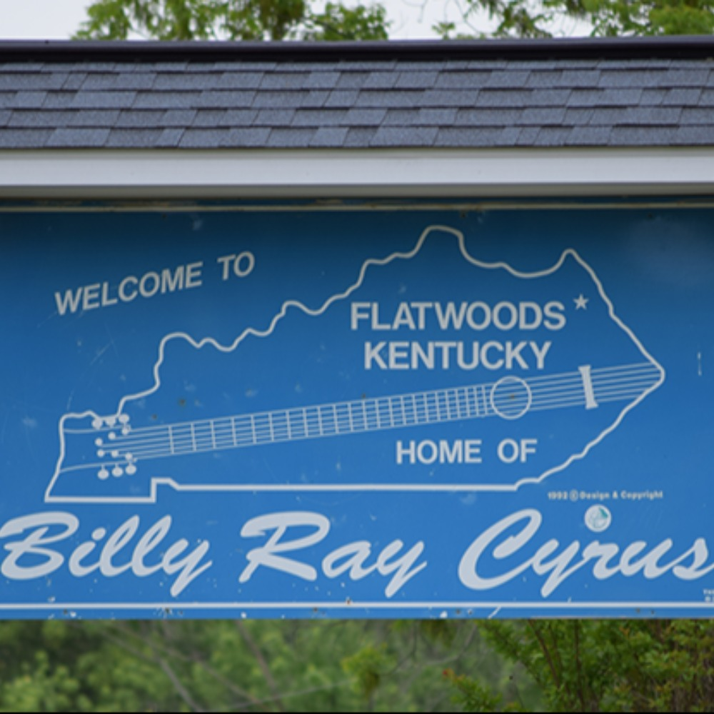 Blue roadside sign with a guitar-shaped state outline reading “Welcome to Flatwoods, Kentucky – Home of Billy Ray Cyrus,” honoring the country music singer’s hometown.