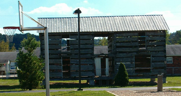 Gilley Log House prior to restoration, showing a deteriorated wooden frame structure with missing walls, situated near a basketball court and lamppost