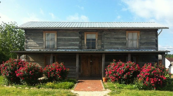 Restored historic Gilley Log House with wooden exterior, front porch, and blooming red bushes on either side of a brick walkway.