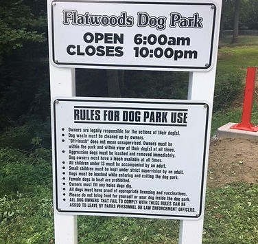 Entrance sign for Flatwoods Dog Park showing operating hours and park rules displayed on a white post.