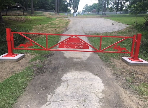 Bright red entry gate labeled 'Flatwoods Dog Park' at the start of a gravel path leading to the fenced park area.