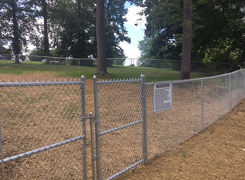 Chain-link fenced area of Flatwoods Dog Park with a gated entrance and posted rules sign, surrounded by trees and open grass.