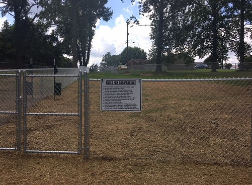Flatwoods Dog Park fence with a visible sign listing usage rules; grassy area enclosed by trees and suburban homes in the background.