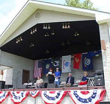 Outdoor stage at B.F. Crager Community Park with patriotic bunting and military flags, where community members are speaking during a public event.