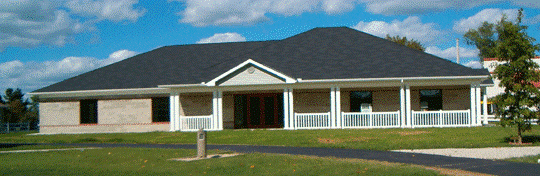 Exterior view of the Flatwoods Senior Center, a single-story building with a dark gable roof, wide front porch, and white columns, set against a blue sky with scattered clouds.