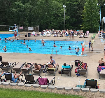 Outdoor swimming pool with people swimming and lounging on chairs, surrounded by trees and greenery.