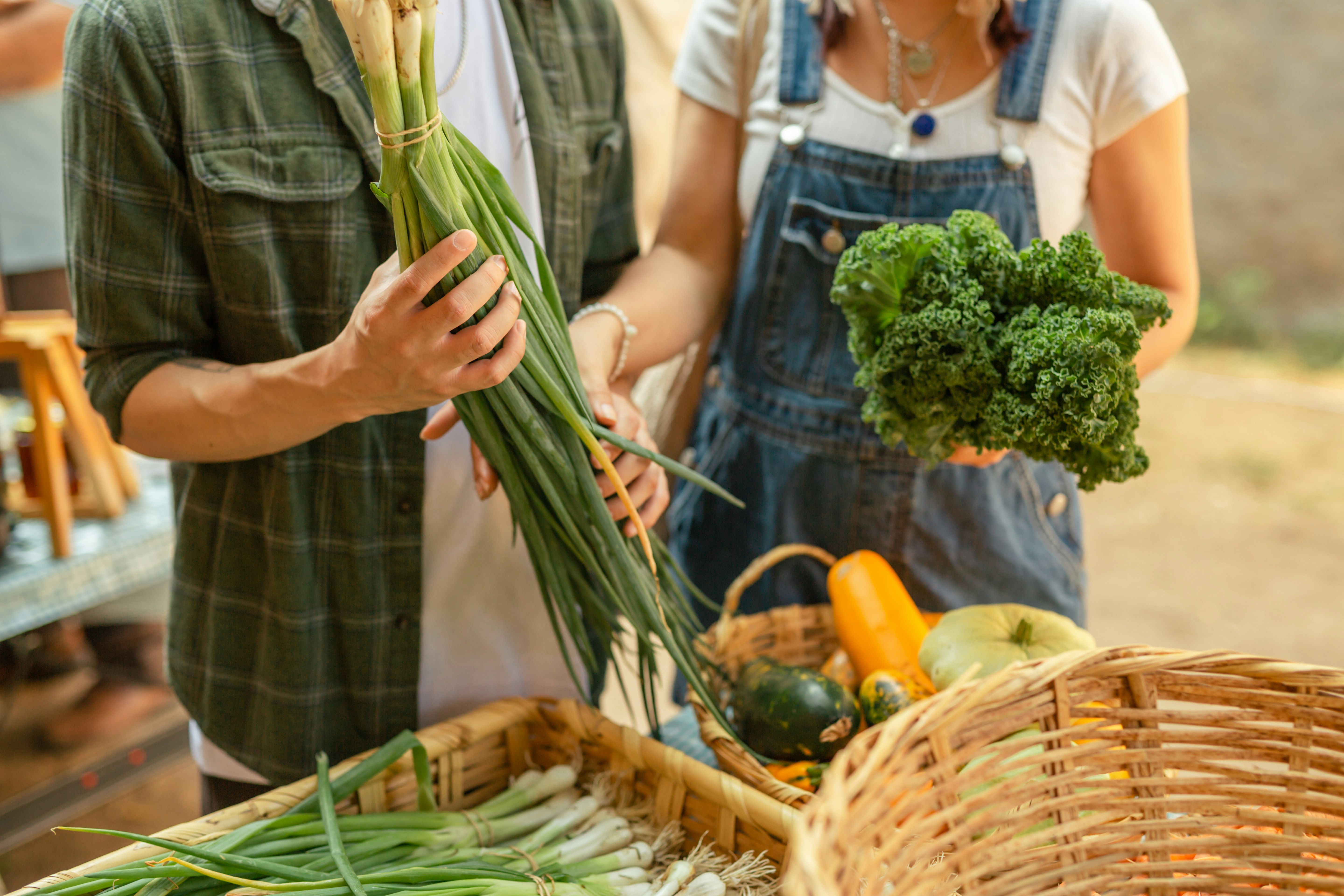 Two people at a farmer’s market holding fresh green onions and kale, standing beside baskets of assorted vegetables.