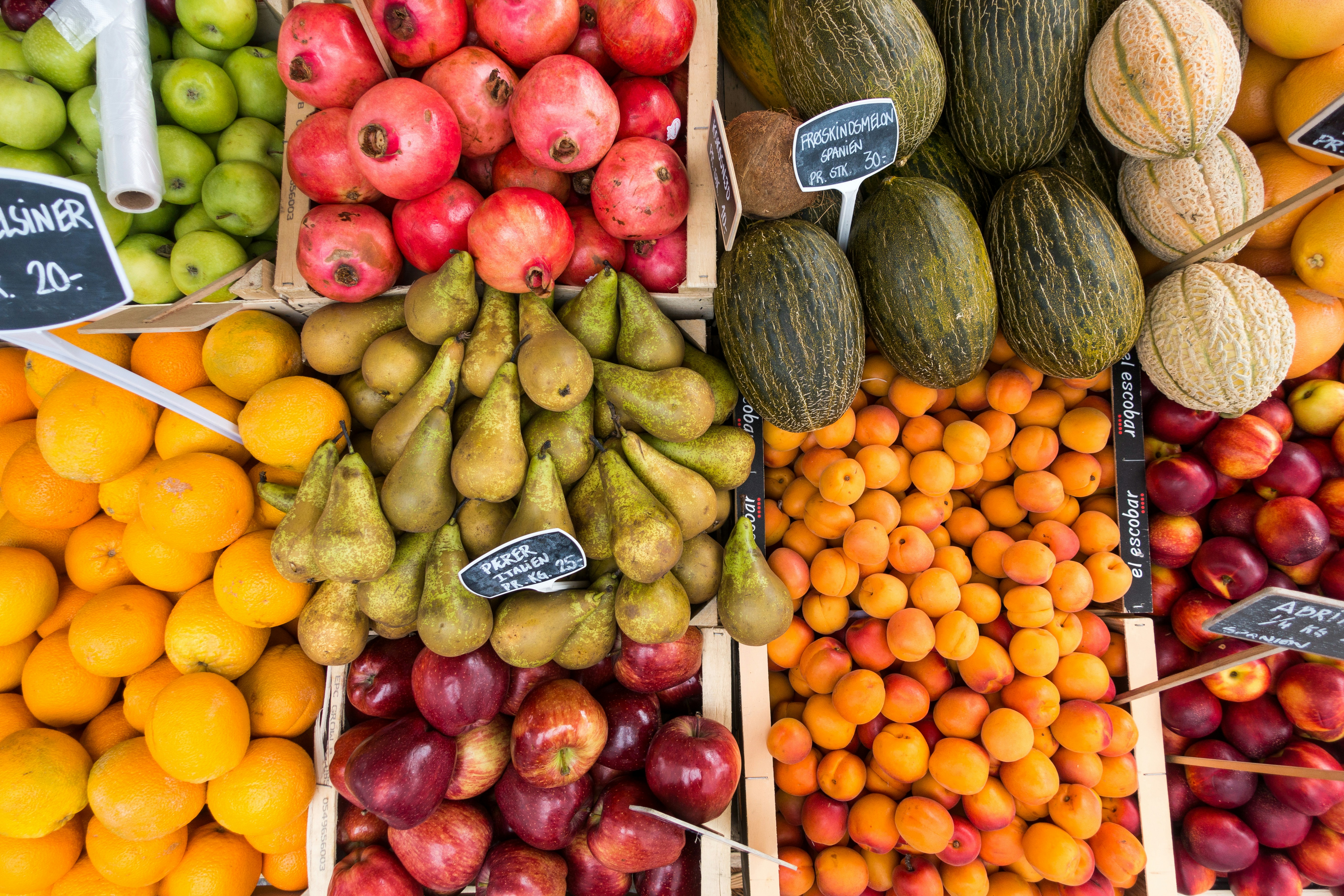 Top view of assorted fresh fruits at a market, including apples, pomegranates, pears, melons, oranges, and apricots arranged in wooden crates.”