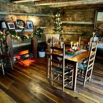 Interior of a rustic log cabin decorated for Christmas with garland, trees, candles, and a wooden dining table and chairs.