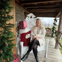 Woman sitting on a white decorative chair on a cabin porch, beside a stack of wrapped gifts and a decorated Christmas tree.