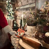 Santa’s hand placing a Christmas book on a decorated rustic table with a rolling pin, candle, and festive greenery in a log cabin setting.
