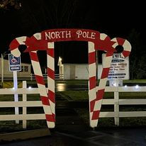 Red and white striped candy cane arch labeled ‘North Pole’ lit up at night near a white fence and signs.