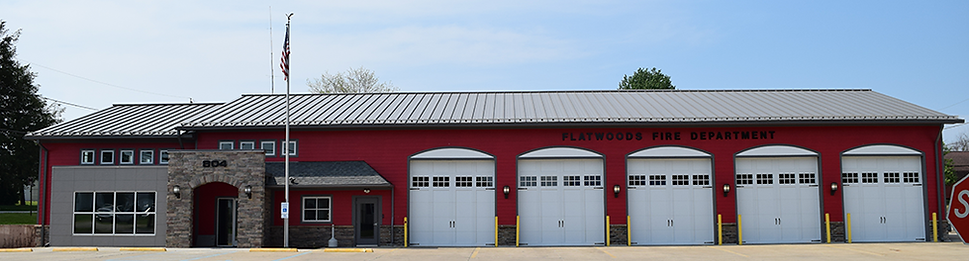 Exterior view of the Flatwoods Fire Department building with five garage bays, a flagpole, and a red and gray facade.