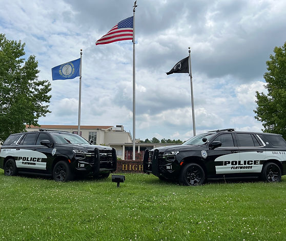 Two Flatwoods Police SUVs parked on grass in front of flagpoles and a high school building, with American, Kentucky, and POW/MIA flags flying.