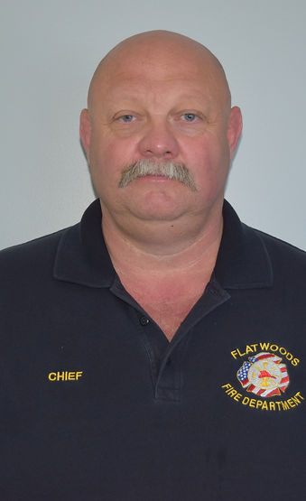 Portrait of a bald, middle-aged man with a mustache wearing a black polo shirt. The shirt has embroidered text reading 'CHIEF' on one side and 'FLATWOODS FIRE DEPARTMENT' along with a fire department emblem on the other side.