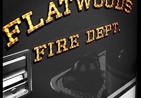 A close-up view of a fire engine displaying the words 'FLATWOODS FIRE DEPT.' in gold and black lettering on a glossy black surface. Reflections in the paint reveal parts of a helmet and surrounding fire equipment.