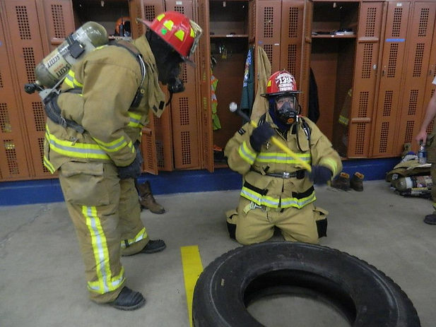 Two firefighters wearing full protective gear and breathing apparatus are participating in an indoor training drill. One firefighter is kneeling in front of a large tire with a tool raised, simulating a striking motion, while the other stands nearby observing. Behind them are open wooden lockers and gear.