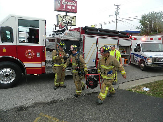 Four firefighters in full turnout gear walk beside a red fire engine parked near a restaurant sign that reads 'Giovanni’s Pizza.' An EMS vehicle and an individual in a yellow vest are visible in the background. The scene takes place on a paved roadside during daylight.