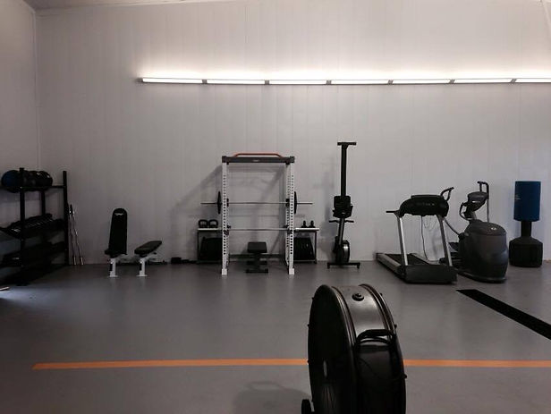 A clean, well-lit fitness room with a variety of workout equipment including free weights, a bench, a squat rack, a rowing machine, a treadmill, and an elliptical trainer. The room features a smooth gray floor and a long overhead light fixture on the white wall.