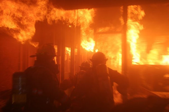 Three firefighters in full turnout gear and self-contained breathing apparatus advance through a burning structure engulfed in flames. The interior is filled with intense orange fire and thick smoke, with flames covering walls and ceiling beams. The scene conveys urgency and danger within a fully involved fire