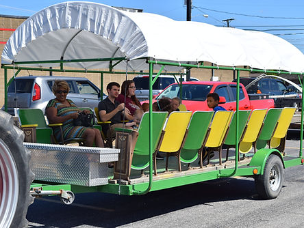 A green and yellow covered wagon pulled by a tractor carries passengers of various ages, sitting on molded seats, along a paved road in a parking lot setting.