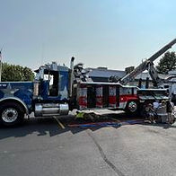 A large patriotic-themed tow truck with chrome accents is parked in a lot, with families and children gathered around to view it during a sunny day at a community truck event.