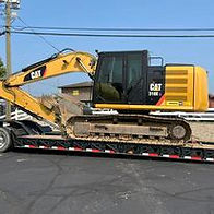 A large yellow Caterpillar 314E excavator rests on a black trailer in a parking lot, displayed for visitors to examine up close during a public vehicle showcase.