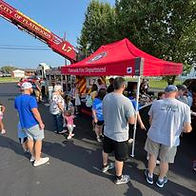 Families visit a red canopy booth labeled “Flatwoods Fire Department” with a ladder truck parked behind it. People are interacting with firefighters and exploring fire safety materials.