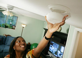 A woman reaches up to test or install a smoke alarm on the ceiling in her living room. A ceiling fan, sofa, and lamp are visible in the background.
