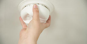 A close-up of a hand installing or testing a white smoke alarm on a ceiling.