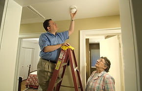 A man standing on a ladder tests or installs a ceiling-mounted smoke alarm while a woman watches from the floor in a hallway of a home.