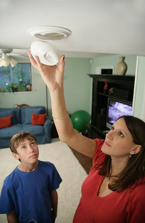 A woman adjusts or tests a smoke alarm on the ceiling while a young boy looks on from below in a living room with a sofa and television in the background.