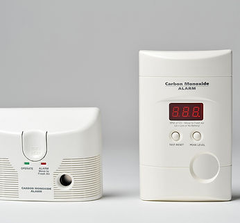 Two rectangular carbon monoxide alarms: the left unit has green and red status lights, a test button, and a speaker grill; the right unit displays a digital reading of “888” on a red LED screen and has three buttons labeled “Test/Reset,” “Peak Level,” and a large circular control at the bottom.
