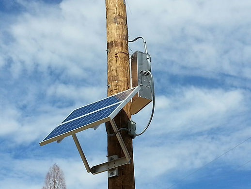 Solar-powered equipment mounted on a wooden utility pole under a partly cloudy sky. The setup includes a tilted solar panel, a metal enclosure, and visible wiring, likely used for powering remote communication or monitoring devices.