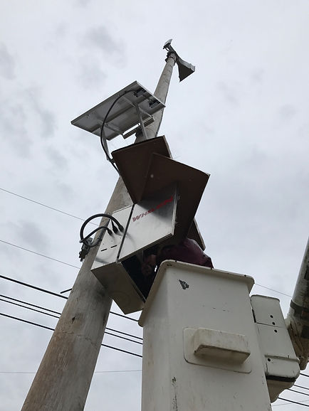 Upward view of a tall utility pole equipped with a solar panel, a metal enclosure box, and a small weather or communication device mounted at the top. A worker in a bucket lift is servicing the equipment. Several power lines run horizontally across the background under an overcast sky.