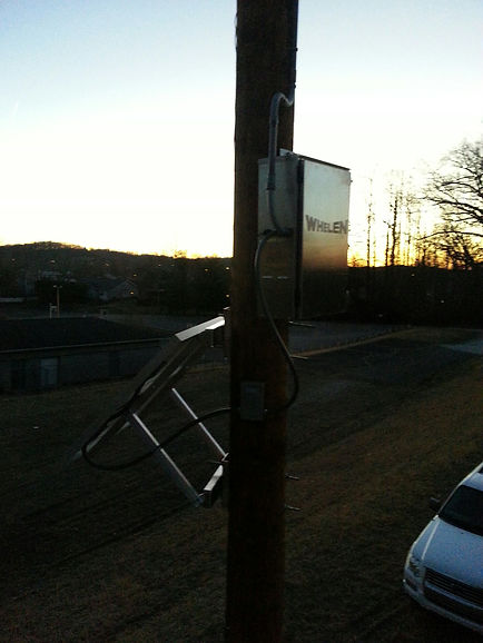 Solar-powered equipment mounted on a wooden utility pole at sunset. The setup includes a tilted metal frame for a solar panel and a metal control box labeled “Whelen.” Power cables run from the box along the pole. A white vehicle and grassy area are visible below, with silhouettes of trees and buildings in the background.