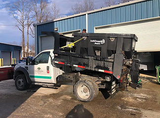 A white maintenance truck fitted with a large black salt spreader is parked outside a blue maintenance building with open garage doors. The ground appears damp and muddy.