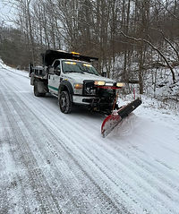A white snow plow truck is clearing a snow-covered rural road bordered by trees in winter. The truck