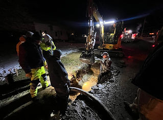 A group of workers in reflective gear perform nighttime utility work using a yellow excavator to dig in a street. The scene is brightly lit, and steam or smoke is visible in the cold air.
