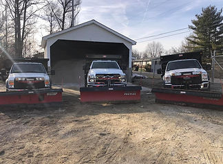 Three white snow plow trucks are parked side by side, facing the camera in front of a large garage. The trucks are equipped with wide red plows.