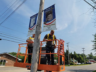 Two utility workers stand in an orange lift, installing commemorative banners on a wooden utility pole along a roadside. The banners honor a local individual, and a clear blue sky is visible in the background.