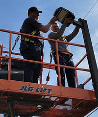 Two workers in safety harnesses stand in an orange lift, installing or repairing a streetlight fixture on a utility pole during the day.