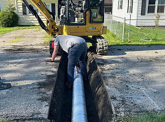 A worker bends over in a freshly dug trench, guiding a large corrugated drainage pipe while a yellow mini excavator is positioned nearby in a residential area.