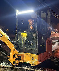A worker operates a yellow compact excavator at night, illuminated by bright lights atop the cab. Some snow is visible on the ground.