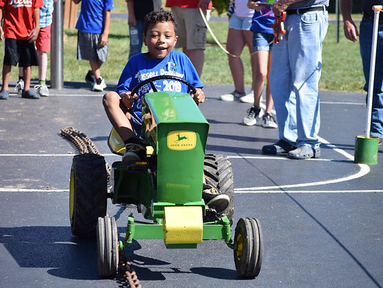 A young boy rides a small green John Deere pedal tractor on a blacktop track while spectators watch and cheer.