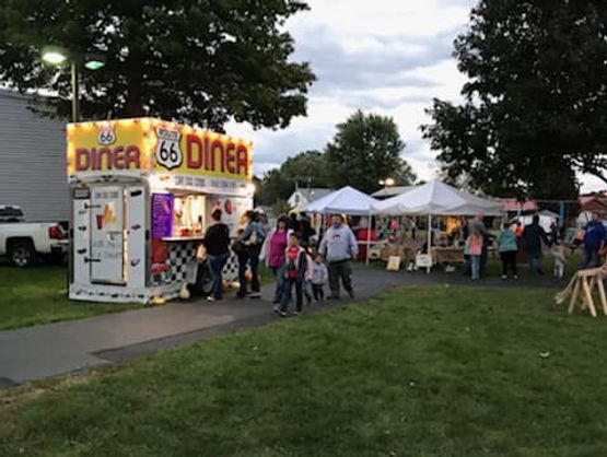 Families walk past a brightly lit Route 66 Diner food stand and white vendor tents at an outdoor evening festival.