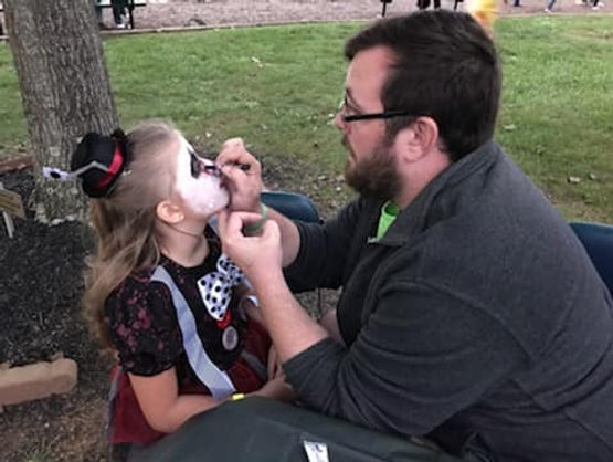A man applies face paint to a young girl dressed in a Halloween costume, sitting outdoors at a table under a tree.